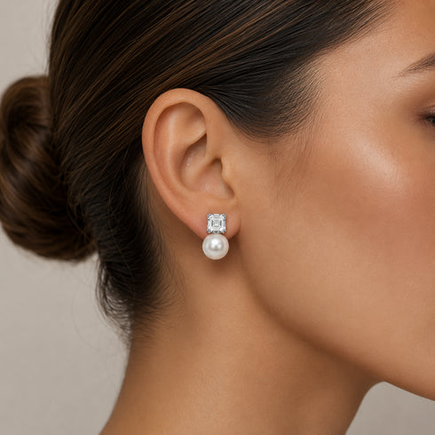 Silver earrings with a square clear gemstone above a dangling white pearl.