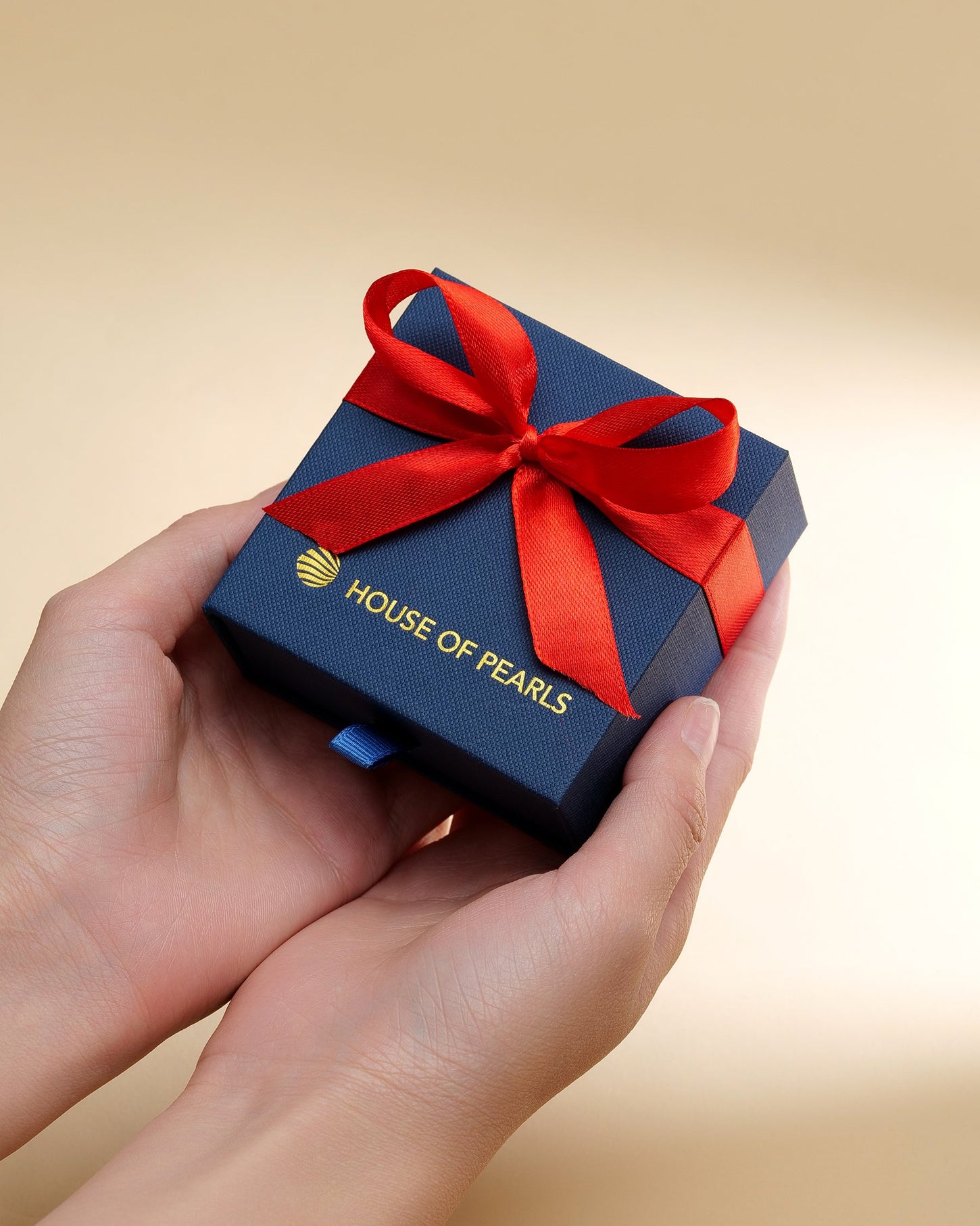 Hand holding a blue gift box with a red ribbon featuring 'House of Pearls' on a beige background.