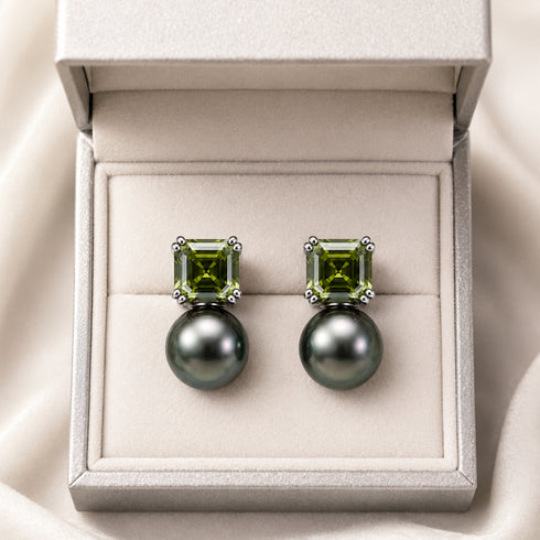 Pair of silver earrings with square olive-green gemstones above round Tahitian black pearls in a beige box.